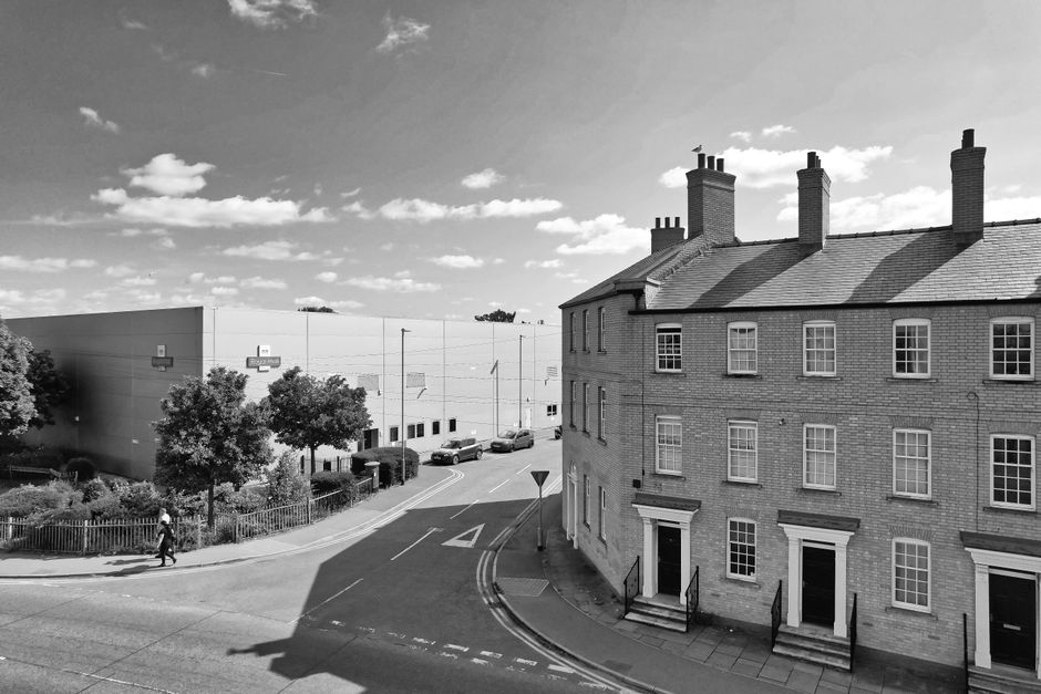 Black and white photograph showing a narrow residential street in Watford, featuring a three-storey brick house with multiple chimneys on the right side, with windows and front doors opening onto the 